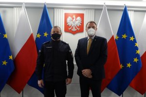 Umundurowany oficer Policji i mężczyzna w garniturze pozują do zdjęcia. Za nimi znajdują się flagi Polski i Unii Europejskiej oraz wisi godło Polski