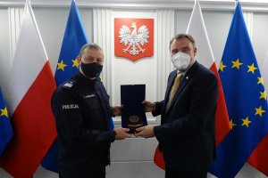 Umundurowany oficer Policji i stojący przy nim mężczyzna w garniturze trzymają pamiątkową tabliczkę. Obaj mężczyźni mają maseczki na twarzach. Za nimi znajdują się flagi Polski i Unii Europejskiej i wisi godło Polski