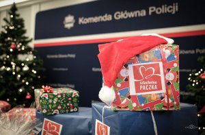 Zapakowany prezent z naklejką szlachetna paczka. Na nim leży czapka Mikołaja. Z tyłu widać przystrojoną choinkę i inne prezenty, a na ściance napis Komenda Główna Policji