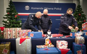 trzej umundurowani oficerowie policji - Komendant Główny wraz z zastępcami - pakują prezenty w ramach akcji szlachetna paczka. Przed nimi i z boku leżą zapakowane prezenty, a z lewej i z prawej strony widać stojące dwie przystrojone choinki. Nad głowami mężczyzny, z tyłu na ściance widoczny jest napis Komenda Główna Policji