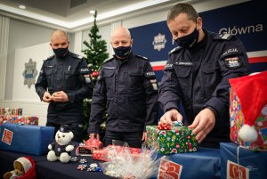 trzej umundurowani oficerowie policji - Komendant Główny wraz z zastępcami - pakują prezenty w ramach akcji szlachetna paczka. Przed nimi i z boku leżą zapakowane prezenty, a z lewej i z prawej strony widać stojące dwie przystrojone choinki. Nad głowami mężczyzny, z tyłu na ściance widoczny jest napis Komenda Główna Policji