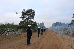 umundurowani policjanci z tarczami i w białych kaskach stoją wzdłuż drogi na granicy. Przed nimi widać drut kolczasty, nad nimi lata policyjny śmigłowiec