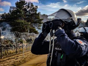 stojący na granicy umundurowany policjant obserwuje okolice przez lornetkę za jego plecami widać drogę i drut kolczasty