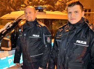 Dwóch umundurowanych policjantów stoi przed radiowozem, pozują do zdjęcia, pada śnieg