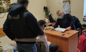 Policjant siedzi za biurkiem i przesłuchuje podejrzanego siedzącego przed nim na krześle. Za plecami siedzącego mężczyzny stoi policjant, który ma na plecach napis Policja