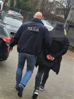 policjant w kurtce z napisem Policja na plecach prowadzi zatrzymanego - widok z tyłu