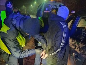 policjant zakłada kajdanki mężczyźnie, z boku stoją dwaj inni funkcjonariusze, a za nimi widać policyjny furgon