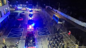 na zdjęciu widać strażaków i policjantów grających na instrumentach wzdłuż szpitala na chodniku, widać niebieska światła i szpaler samochodów służb mundurowych