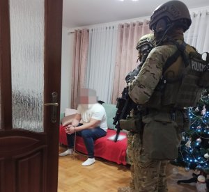 Zatrzymany mężczyzna, który siedzi na kanapie w mieszkaniu i stojący nad nim policjanci