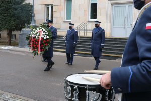 Komendant Główny Policji wraz z dwoma zastępcami. Przed nimi umundurowany funkcjonariusz niesie wieniec z biało - czerwonych kwiatów. Z prawej strony widoczny jest fragment werbla, w który pałeczkami uderza policjant