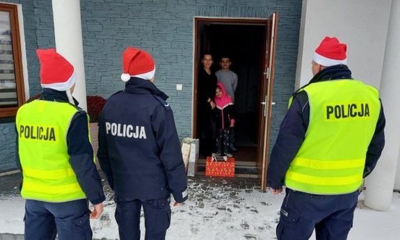 trzej policjanci przed budynkiem z prezentami, w budynku otwarte drzwi, w którym stoją kobieta i dwoje dzieci