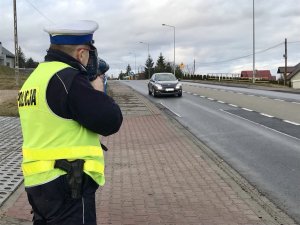 policjant ruchu drogowego trzyma w ręku ręczny miernik prędkości i mierzy prędkość jadącego po jezdni pojazdu