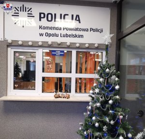 okienko dyżurki Komendy Powiatowej Policji w Opolu Lubelskim. Przed okienkiem widać ustrojoną bombkami i łańcuchami choinkę. na parapecie stoi pociąg