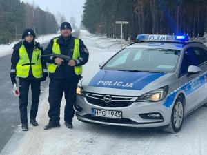 Policjantka i policjant ruchu drogowego obok radiowozu oznakowanego. Mężczyzna posiada broń automatyczną