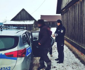 Zdjęcie na zewnątrz. Policjant stoi przy radiowozie, z którego wychodzi starszy mężczyzna. W tle widać kobietę i zabudowania gospodarcze