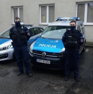 dwaj policjanci stoją przed radiowozem policyjnym, w tle budynek