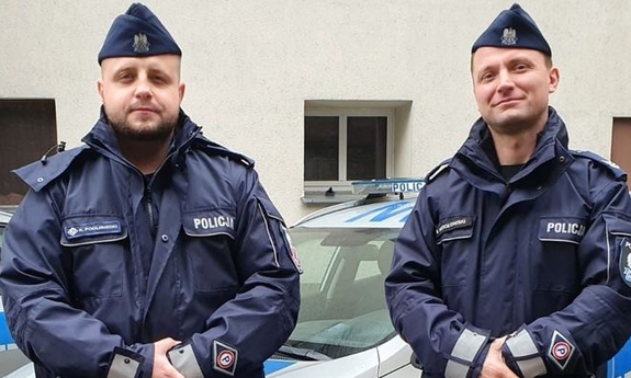 dwaj policjanci stoją przed radiowozem policyjnym w tle budynek