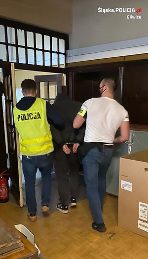 Dwaj policjanci po cywilnemu prowadzą korytarzem w komisariacie zatrzymanego mężczyznę