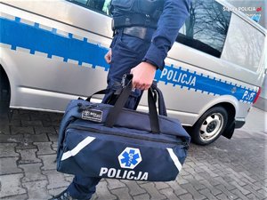 Zdjęcie kolorowe: umundurowany policjant z torbą pierwszej pomocy na tle oznakowanego radiowozu