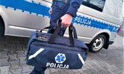 Zdjęcie kolorowe: umundurowany policjant z torbą pierwszej pomocy na tle oznakowanego radiowozu