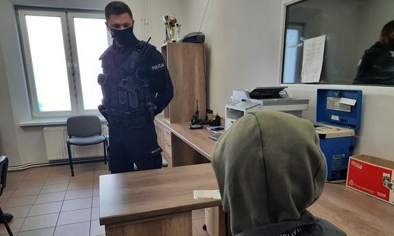 wnętrze komisariatu, policjant stoi naprzeciwko zatrzymanego siedzącego na krześle