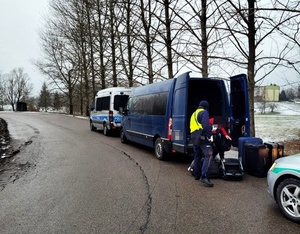 Policyjny radiowóz, za nim niebieski bus z otwarty,i drzwiami, obok ludzie z walizkami, samochód Straży Granicznej