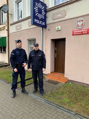 dwóch umundurowanych policjantów, który eskortowali ciężarną kobietę do szpitala stoi przed budynkiem Posterunku Policji