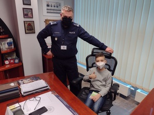 chłopiec siedzi na krześle przy biurku, obok stoi umundurowany policjant