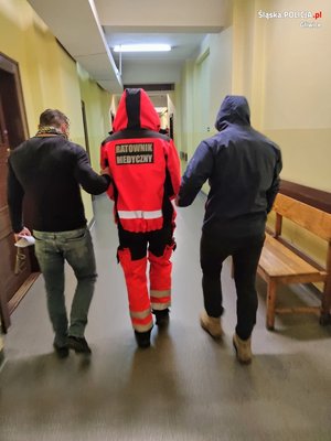 Dwaj policjanci po cywilnemu prowadzą mężczyznę przebranego za ratownika medycznego