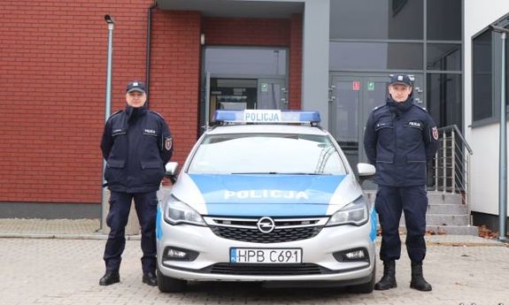dwaj umundurowani policjanci stoją przed budynkiem komisariatu przy radiowozie