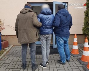 zatrzymany mężczyzna prowadzony do budynku przez dwóch nieumundurowanych policjantów, prowadzących go z dwóch stron pod ramię