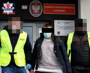 dwóch funkcjonariuszy prowadzi zatrzymanego mężczyznę, w tle budynek komendy powiatowej policji