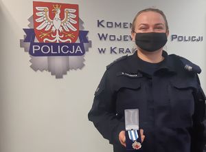 Umundurowana policjantka w maseczce pozuje do zdjęcia na tle ścianki z napisem Komenda Wojewódzka Policji w Krakowie. Policjantka w dłoni trzyma odznaczenie