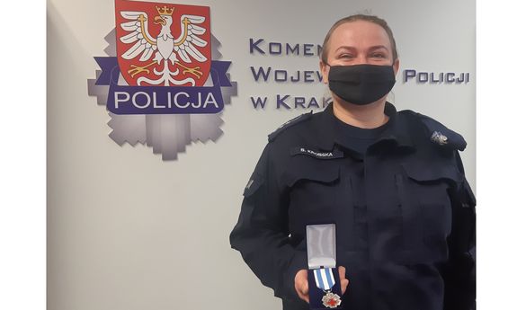 Umundurowana policjantka w maseczce pozuje do zdjęcia na tle ścianki z napisem Komenda Wojewódzka Policji w Krakowie. Policjantka w dłoni trzyma odznaczenie