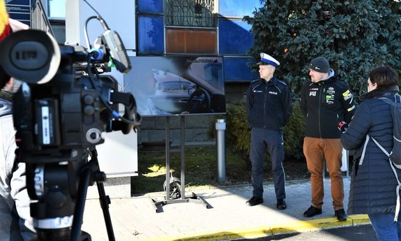 umundurowany policjant stoi obok mężczyzny, na telewizorze wyświetlany jest spot profilaktyczny