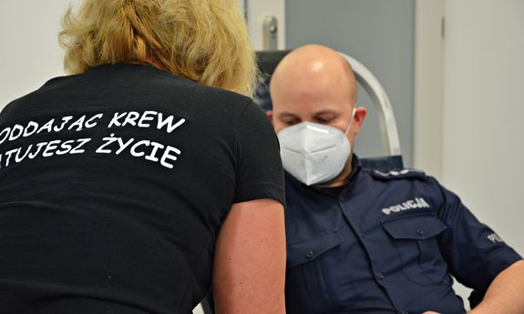 policjant oddaje krew, nad nim stoi kobieta w czarnej koszulce z białym napisem na plecach oddając krew ratujesz życie