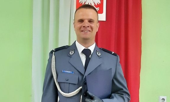 policjant w galowym mundurze pozuje do zdjęcia na tle zawieszonej za nim biało czerwonej flagi i godła Polski