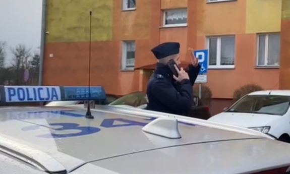 umundurowany policjant stoi przy radiowozie i trzyma telefon komórkowy przy uchu, w tle blok mieszkalny