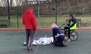 policjantka udziela pomocy chłopcu który zasłabł na boisku, po prawej chłopiec na rowerze, po lewej chłopiec w czerwonej kurtce