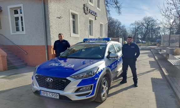 dwóch umundurowanych policjantów stoi przy radiowozie, w tle budynek Policji