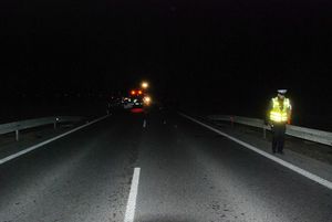 Na zdjęciu - pora nocna, jezdnia, po prawej stronie policjant w odblaskowej kamizelce. W tle widać rozbłyski świateł pojazdów zabezpieczających miejsce wypadku