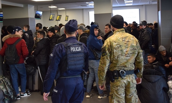 policjanci w mundurach obserwujący liczną grupę pasażerów czekających w kolejce, w terminalu lotniska