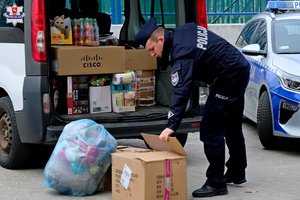 umundurowany policjant pochyla się nad kartonem stojącym na ziemi przed wypełnionym darami samochodem