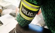 widok na opaskę z napisem policja, którą policjantka pochylona nad zabezpieczonymi ubraniami popakowanymi w worki ma na lewym przedramieniu