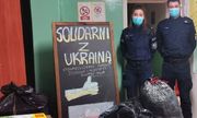 dwoje policjantów biorących udział w zbiórce w pomieszczeniu ze zgromadzonymi przedmiotami, przy tablicy z napisem: Solidarni z Ukrainą