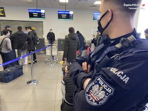 policjant z naszywką na lewym ramieniu z napisem komenda powiatowa policji obserwuje osoby w kolejce do odprawy, w terminalu lotniska
