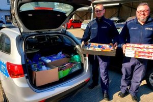 policjanci w mundurach stoją przy radiowozie trzymając w rekach pudełka z produktami, bagażnik wypełniony produktami spożywczymi i chemicznymi