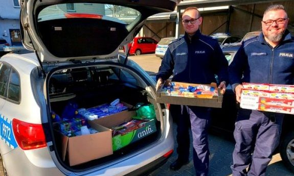 policjanci w mundurach stoją przy radiowozie trzymając w rekach pudełka z produktami, bagażnik wypełniony produktami spożywczymi i chemicznymi