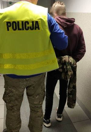 policjant w żółtej kamizelce z napisem policja prowadzi zatrzymanego mężczyznę który był poszukiwany, zatrzymany trzyma ręce spięte w kajdanki z tyłu