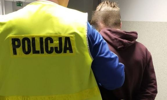 policjant w żółtej kamizelce z napisem policja prowadzi zatrzymanego mężczyznę który był poszukiwany, zatrzymany trzyma ręce spięte w kajdanki z tyłu